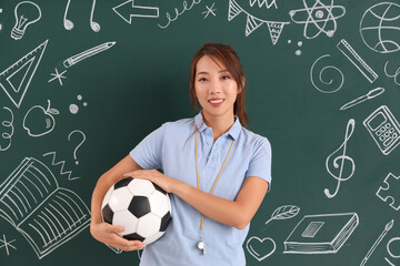 Wall Mural - Female Asian PE teacher with soccer ball near blackboard in classroom