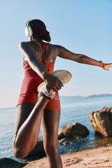 Wall Mural - Young woman stretching before running by the seaside, wearing headphones and athletic gear, enjoying a bright sunny day