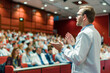 © Nelly - A doctor in a white coat delivers a lecture to a large audience in a modern auditorium, engaging attendees in a medical conference or seminar