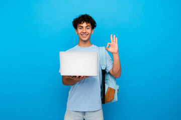Young teenager high school boy guy holding laptop computer showing okay gesture while using it for surfing webpages on Internet, social media posting,e-learning remotely isolated in blue background