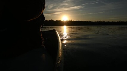 Wall Mural - View looking backwards as a canoe is being paddled along a large lake as the sun is setting over an evergreen forest. The canoe and the person paddling fill the left side of the frame.
