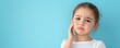 © WE.ARE.AI - Little girl with a toothache touching her cheek, standing against a blue background, expressing pain and discomfort.
