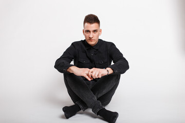 Stylish handsome young man model in fashionable black shirt with jeans sitting on white background in studio