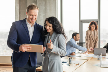 Aufkleber - Two business professionals are working together in a modern office setting. They are standing near a large wooden table with other colleagues in the background. The man is holding a tablet
