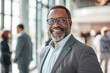 © GraysonStock - A middle-aged African-American businessman standing in an office, smiling and looking at the camera, dressed smartly in a gray suit.