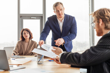 Poster - A businessman in a blue suit stands and hands documents to a seated colleague while another sits in the background, all gathered around a conference table in an office setting.
