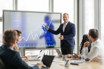 Aufkleber - A businessman in a suit stands in front of a large screen, pointing to an upward trending graph. He is presenting to a group of colleagues who are seated at a conference table, listening attentively