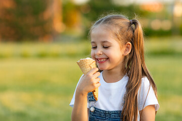 A happy laughing child in summer clothes eats ice cream on a hot day in park. Ice cream in a waffle cone. A happy and contented child at summer.