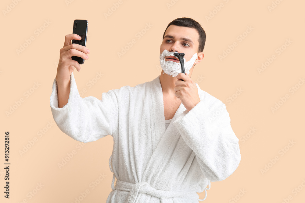 Handsome young man with razor shaving his face and taking selfie on beige background