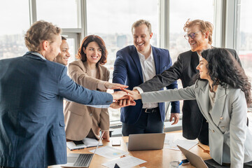 Aufkleber - A diverse business team of six people stand around a table with laptops and paperwork in a modern office. They have their hands stacked in the center, smiling and celebrating a shared success.