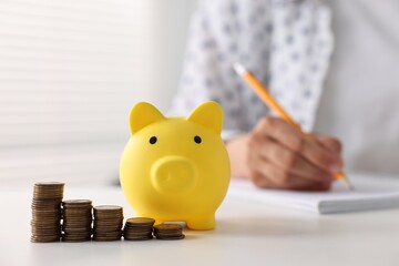 Poster - Woman taking notes at table indoors, focus on piggy bank and coins