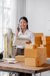 © David - Smiling happy young Asian woman sme entrepreneur receiving phone call for new sales order among boxes of product with laptop computer. Success online business and sme online concept.