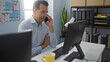 © Krakenimages.com - Hispanic man talking on a mobile phone in an office, looking at a computer monitor with a serious expression, surrounded by organized folders and decor in a well-lit environment.