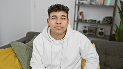 Wall Mural - A handsome young man wearing a white hoodie comfortably seated on a grey couch indoors, exuding a casual, relaxed atmosphere.