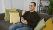 © Krakenimages.com - Cheerful middle-aged man texting on smartphone, sitting on a gray couch surrounded by colorful pillows in a cozy living room.