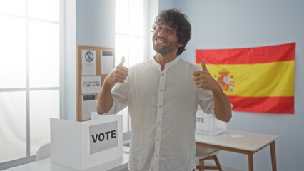 Young man in an electoral room in spain gives thumbs up in front of a voting booth and spanish flag, expressing happiness and confidence in the voting process.