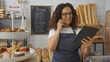© Krakenimages.com - Woman working in a bakery, holding a tablet and talking on the phone, in front of shelves with bread and pastries, wearing an apron, with glasses and curly hair.