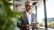 © halo - A smiling businessman in his forties holding an iPad, dressed smartly and wearing glasses with gray hair, stands on the balcony of a modern office space.