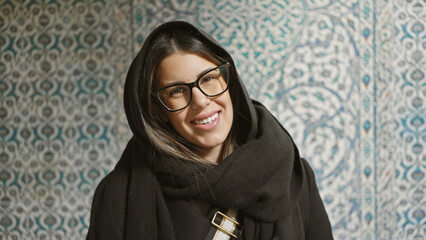 Wall Mural - A smiling young woman with glasses stands in front of an ornate turkish tile wall, exuding a sense of joy and cultural immersion.