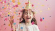 © VK Studio - A joyful girl with a party hat enjoying a shower of colorful confetti, expressing pure happiness against a pastel pink background.
