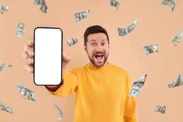 Wall Mural - Happy man showing mobile phone with empty screen under money rain on dark beige background