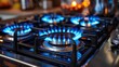 © Tanakorn - Close-up view of a gas stovetop burner with blue flames ignited, showcasing a modern kitchen appliance with a sleek black and stainless steel design.