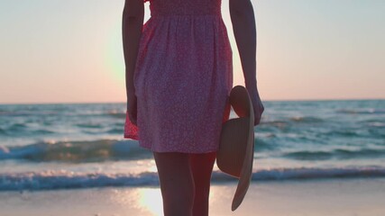 Wall Mural - summer mood. a slender girl in a dress and with a hat in her hands walks along the beach at sunset, enjoying her summer vacation.  a girl in a summer dress walks along the seashore.