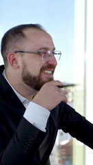 Wall Mural - Smiling business worker talking to someone in office. Portrait of entrepreneur in glasses and costume sitting at his desk on the background of window in the city. Vertical video