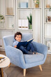 © Iona - Young boy sitting on a light blue armchair, holding an open book, in a cozy, modern living room with shelves in the background