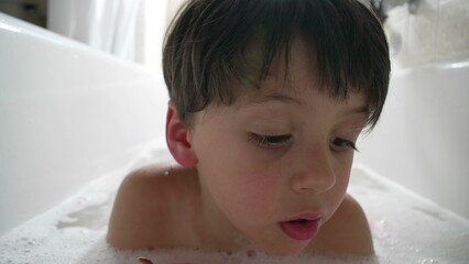 Close-up of a young boy's face as he leans over the bathtub, surrounded by bubbles. His expression is one of curiosity and concentration, enjoying the fun and relaxation of bath time