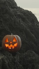 Wall Mural - Vertical video. Jack-O'-Lantern Pumpkin with Flickering Candlelight Inside Stands on Sharp Rocks in the Sea. Halloween Scene.