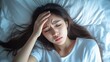 © Plaifah - Unwell young Asian woman lying in bed, holding her forehead in discomfort, suffering from a headache.