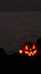 Wall Mural - Vertical video. Peeking Out from Behind the Rocks, Watching the Jack-O'-Lantern Pumpkin Against the Sea at Night. Altar on the Rocks for the Celebration.