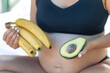 © puhimec - Pregnant woman holding avocado and bananas, health and maternity concept.