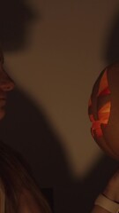 Wall Mural - Vertical video. A Woman in a Dark Room Holds a Jack-O'-Lantern, Staring into Its Eyes. Their Shadows Cast on a White Wall with Burning Candles. A Cozy Halloween Setting.