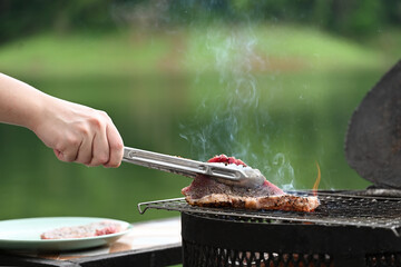 A hand using tongs to flip a seasoned steak on an outdoor grill, with smoke rising and the green scenery in the background, captures the essence of outdoor cooking