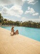 © Wirestock - Woman in a black swimsuit sitting by the edge of a swimming pool with a scenic natural view.