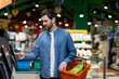 © Liubomir - Man at self-checkout machine in grocery store holding orange basket with vegetables. Concept of modern shopping, technology use, convenience, and customer experience. Shopping with card payment.