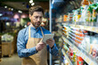 © Liubomir - Grocery store employee with apron using tablet for inventory management in food aisle. Worker checking stock and organizing products in supermarket. Efficient retail operations with technology