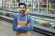 © Liubomir - Supermarket worker standing confidently with arms crossed in front of freezer section. Employee wearing blue shirt and beige apron smiling. Grocery store setting with various frozen products in the