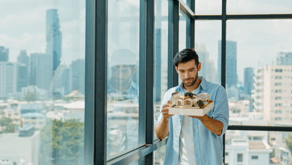 Wall Mural - Businessman in casual outfit holding house model while checking house construction. Architect engineer inspect building model while standing near window with skyscraper. Engineering. Tracery