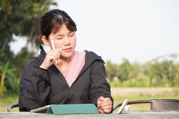 Wall Mural - Young asian woman is thinking while working remotely at outdoor area or park using a tablet. Natural light with trees and natural background. Concept for freelancer.