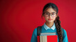 © Vadym Hunko - Serious Asian teenage girl with glasses and a long braided hairstyle, wearing a blue sweater and holding books, standing against a solid red background. The atmosphere is studious and focused.