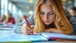 © Vadym Hunko - Focused Caucasian teenage girl with long blonde hair writing in a notebook with a red pen, in a brightly lit classroom. The atmosphere is studious and concentrated.