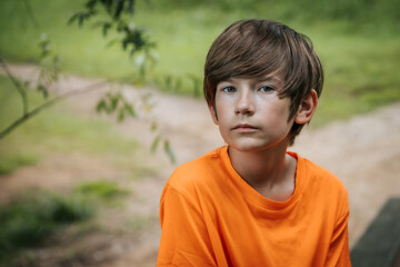 A young boy with a serious expression sits outdoors, wearing an orange shirt. The background features soft greenery, creating a serene and natural setting.