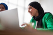 © Studio Marmellata - A woman in a black hijab and green blazer smiles as she works on her laptop in a bright office. Her positive demeanor adds to the professional yet friendly atmosphere of the workspace.