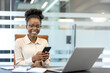 © Liubomir - Young African American businesswoman at workplace inside office smiling at camera holding phone. Professional, confident, and productive. Represents modern business, communication, and technology.