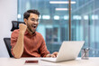 © Liubomir - Young man celebrating successful achievement while working on laptop. Joyful professional feeling accomplished in business environment. Smartphone on desk.