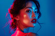 © hocine - A young woman with long dark hair looks directly at the camera, lit with blue and red light, showcasing her makeup and jewelry.