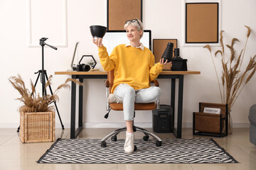 Sticker - Young woman in eyeglasses with photo camera lenses sitting on armchair near workplace with modern computer in stylish office